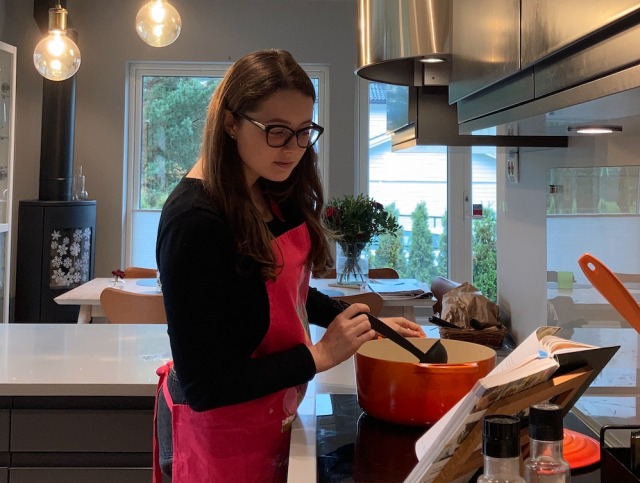 Au pair Mariia Zaichuk står på kjøkkenet og rører i en gryte, mens hun leser i en kokebok som ligger på kjøkkenbenken. 