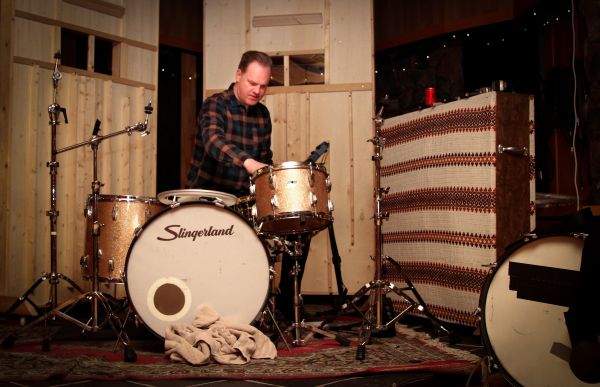 Musikkprodusent Christer Krogh sitter og spiller på et trommesett i studio. Han ser på trommene, og er konsentrert. Han har brunt hår og en rutete skjorte i flere farger. 