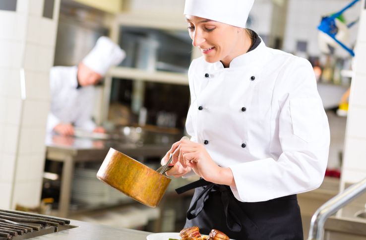 Kvinnelig kokk på et kjøkken, ikledd hvitt kokkeantrekk. Hun holder en kjele i hånden.
