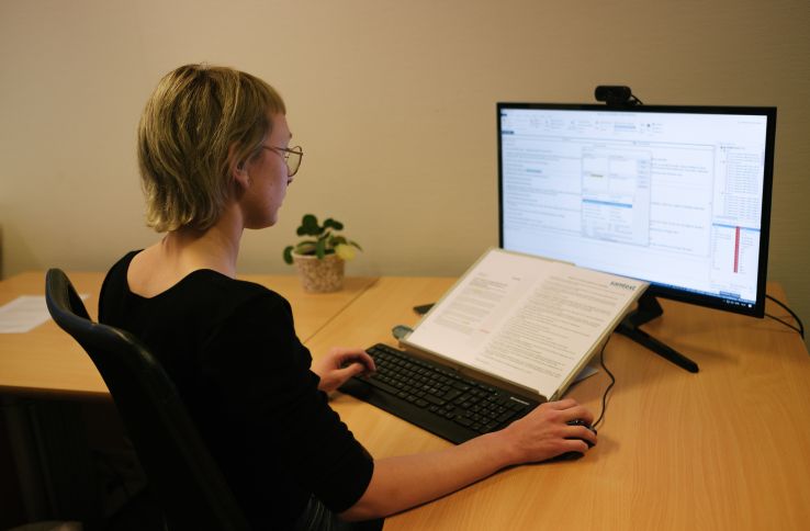 En dame sitter foran en PC, foran skjermen ligger en åpen bok med tekst 