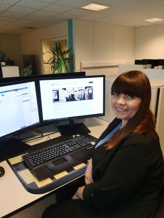 Kvinne i svarte klær sitter foran dataskjermer ved skrivebord og smiler.