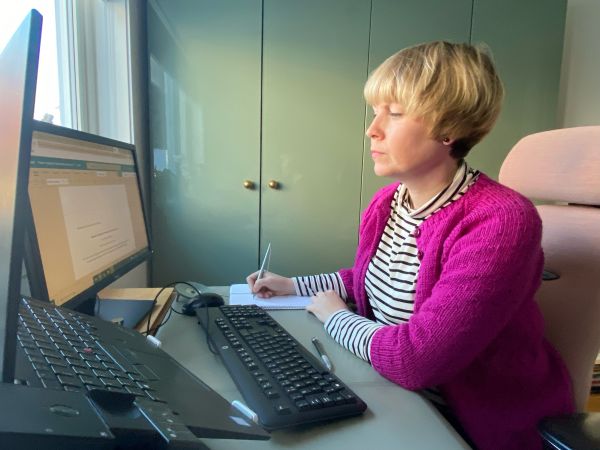 Kvinne med rosa jakke sitter ved skrivebord og skriver på datamaskin
