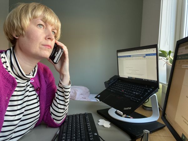 Kvinne med rosa jakke sitter ved skrivebord foran datamaskin og prater i mobil.