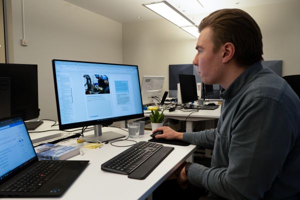 Mann sitter og ser på dataskjermen ved et skrivebord, fremme har han dokumenter.