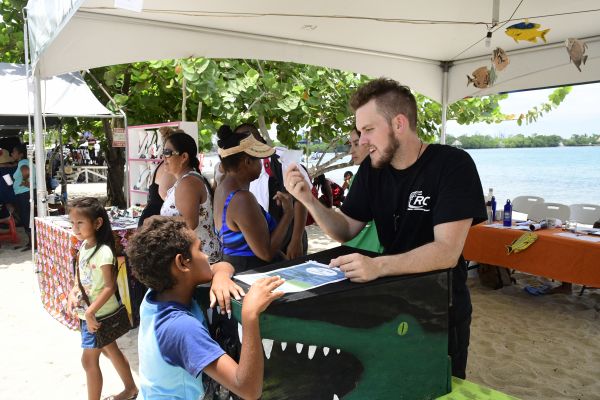 Mann i svart t-skjorte står ved et bord formet som en krokodille og snakker med en gutt i tropiske omgivelser