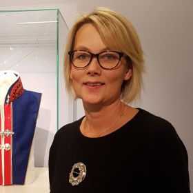 Portrettbilde av duojár Jorunn Løkvold. Hun har på seg briller, en svart overdel og en brosje midt på brystet. Halvveis i bakgrunnen ser vi litt av en blå og rød kofte. 