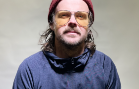 portrettfoto av regissør Carl Christian Lein Størmer, Mann med skjegg, gule briller og rød strikkalue.