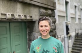Scenograf Thale Kvam Olsen er avbildet foran en sideinngang til Den Nationale Scene i Bergen. Hun er iført grønn genser og mørkt, oppsatt hår. Bak henne ser vi en grønn dør og grått mursteinbygg.
