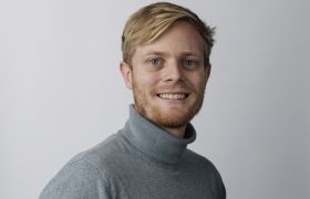 Portrettbilde av solcelleforsker Magnus Moe Nygård. Han står foran en grå bakgrunn og har på en grå genser. 
