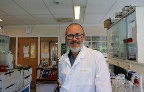 Kjemiker John A. Gjerde er avbildet på laboratoriet der han jobber. Han er avbildet fra livet og opp, og har svarte briller, skjegg og smiler mot kamera. Han er iført hvit labfrakk, og i bakgrunnen ser vi permer og arbeidsredskaper.