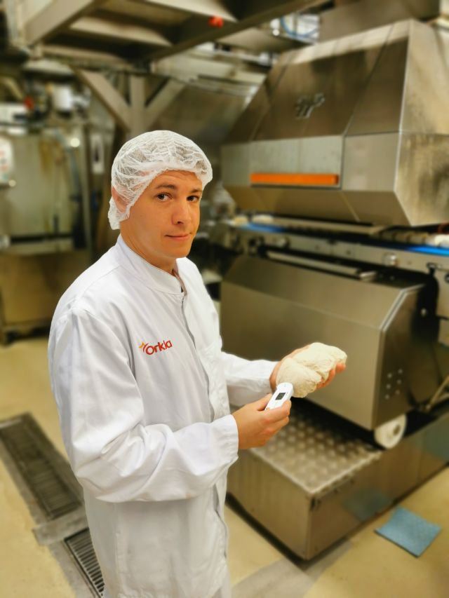 Fagarbeider i industriell matproduksjon Håvard Strandos holder opp en pizzadeig i hendene og ser inn i kamera. Han har på seg hvitt arbeidsantrekk og hårnett og står i bakeriavdelinga på fabrikken
