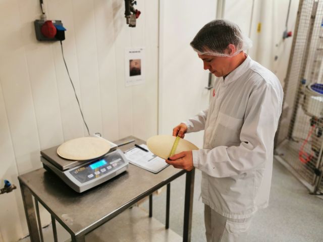 Fagarbeider i industriell matproduksjon Håvard Strandos, står ved et bord og måler opp ferdige pizzabunner med et målebånd. Han har på seg hvitt arbeidsantrekk og hårnett. Det ligger en pizzabunn på vekta på bordet også. 