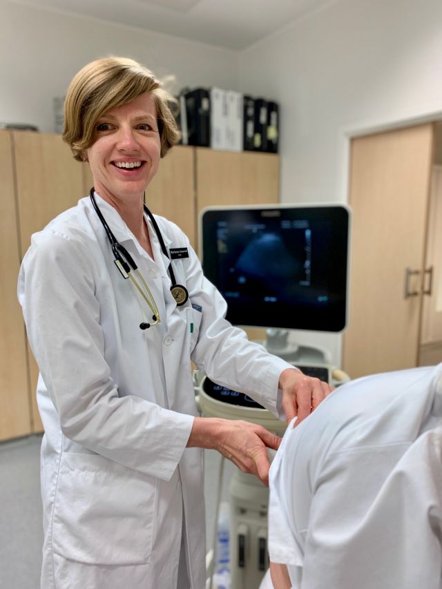 Overlege og førsteamanuensis Marianne Aanerud har på seg hvit arbeidsfrakk og undersøker en pasient mens hun smiler og ser inn i kamera.