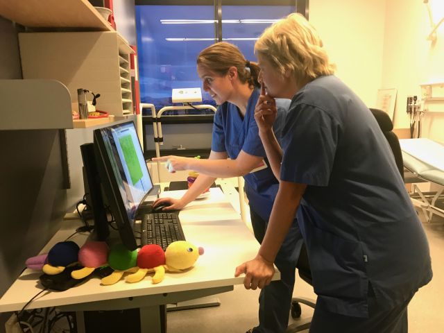Barne- og diabetessykepleier Rigmor Knutsen i dialog med en kollega om noe på dataskjermen. Det ligger en barneleke på skrivepulten. Begge har på seg blå arbeidsuniform.