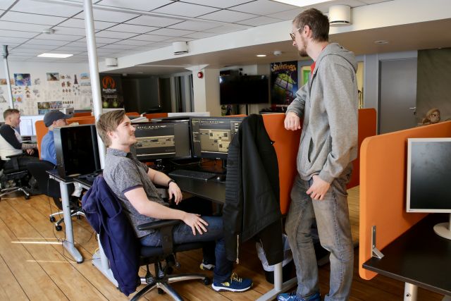 Spelprogrammerar Marcus André Lie sitt ved ein arbeidspult og ser mot kollegaen sin som står ved pulten hans og pratar. 