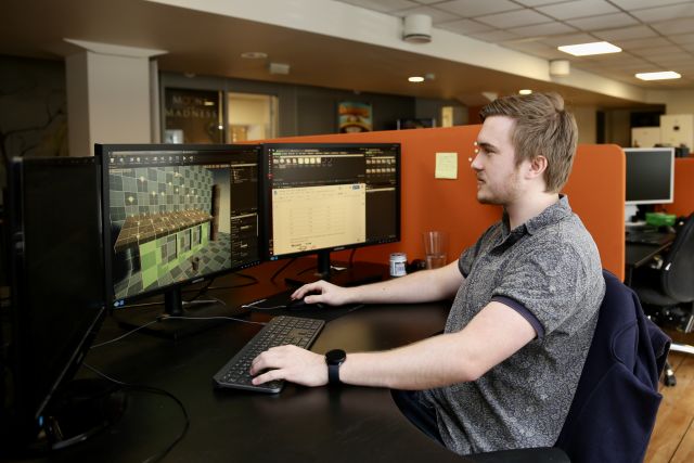 Spelprogrammerar Marcus André Lie sittande ved ein arbeidspult med to skjermar. Han ser mot einl teikning av eit digitalt rom i eit spel.  