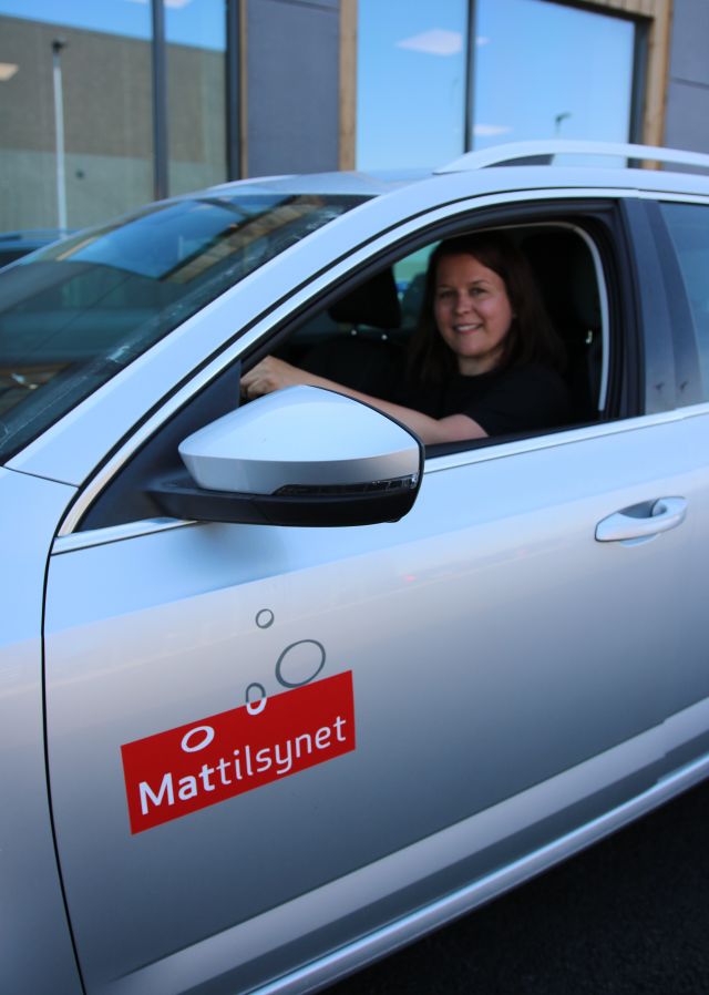 Matteknolog Kari Haugstad er avbildet sittende bak rattet i en grå bil med Mattilsynets logo. Vi ser et bygg i bakgrunnen, og ser deler av bilen på førersiden. Hun har svart T-skjorte og mørkt langt hår.