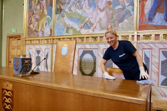 Renholdsoperatør Maricela Naprica er avbildet mens hun rengjør bystyresalen i Haugesund rådhus. Hun tørker over et stort bord, og i bakgrunnen ser i malerier og byvåpenet til Haugesund. Hun har lyst hår satt opp i en knute, og er iført mørkeblå T-skjorte 