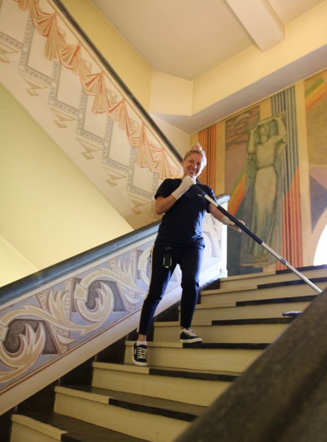 Renholdsoperatør Maricela Naprica er avbildet mens hun rengjør en trapp i Haugesund rådhus. Hun bruker en mopp, og står midt i trappen. Hun er vendt mot kamera og smiler, og er iført mørkeblå T-skjorte og bukse. 