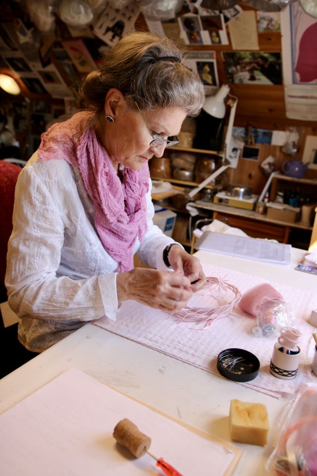 Modist Kirsti Nordberg Moe er avbildet på atelieret sitt på Stord. Hun sitter ved arbeidsbordet, og er iført briller, hvit skjorte og et rosa sjal. Hun har håret løst oppsatt i en knute, og bak henne skimter vi hatter og utstyr. 