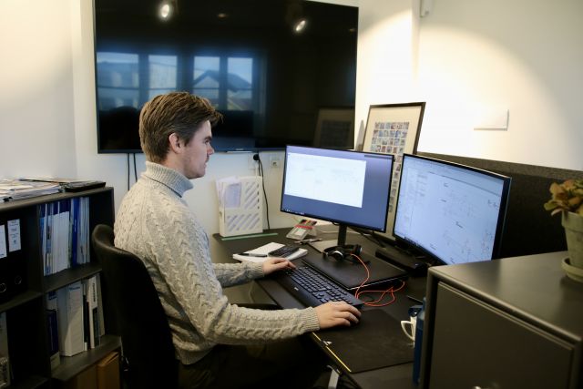 Ingeniør energi og miljø i bygg, Jørgen Knutsen, sitter foran to store pc-skjermer og jobber. Han er iført beige høyhalset genser, og vi ser han fra siden. 