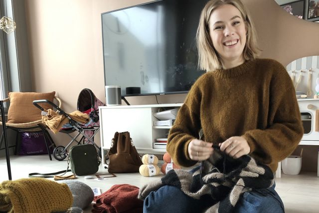 Strikkehåndverker Pia Marlene Øye Amundsen sitter på gulvet hjemme med strikketøyet i hendene og garn, oppskrifter og ferdige strikkegensere ligger rundt henne.