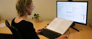 En dame sitter foran en PC, foran skjermen ligger en åpen bok med tekst 