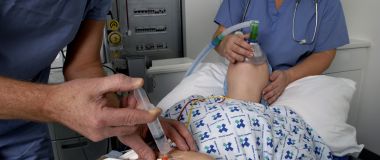 Lege injiserer innsovningsmiddel i hånden til pasient mens en anestesisykepleier bedøver pasienten med oksygenmaske.