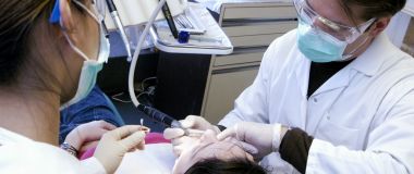 Tannlege polerer tennene til en pasient, mens en tannhelsesekretær holder et beger med slipepasta som brukes i poleringen.