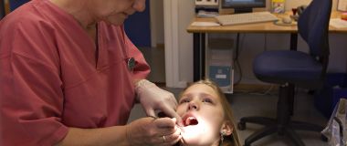 Tannpleier hos skoletannlegen kontrollerer tennene til ei jente. I bakgrunnen er en dataskjerm med røntgenbilde av tenner.