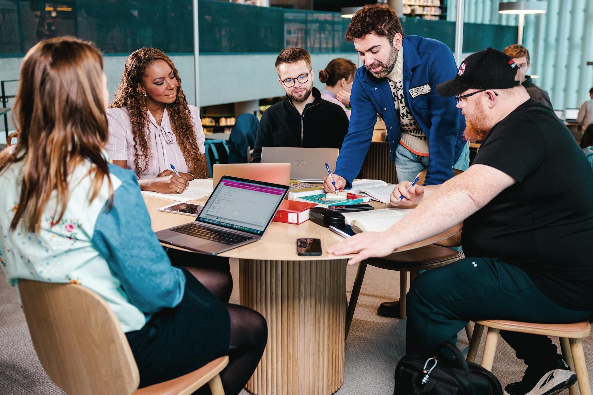 Fem voksne personer sitter og står rundt et bord, og jobber med skolearbeid.